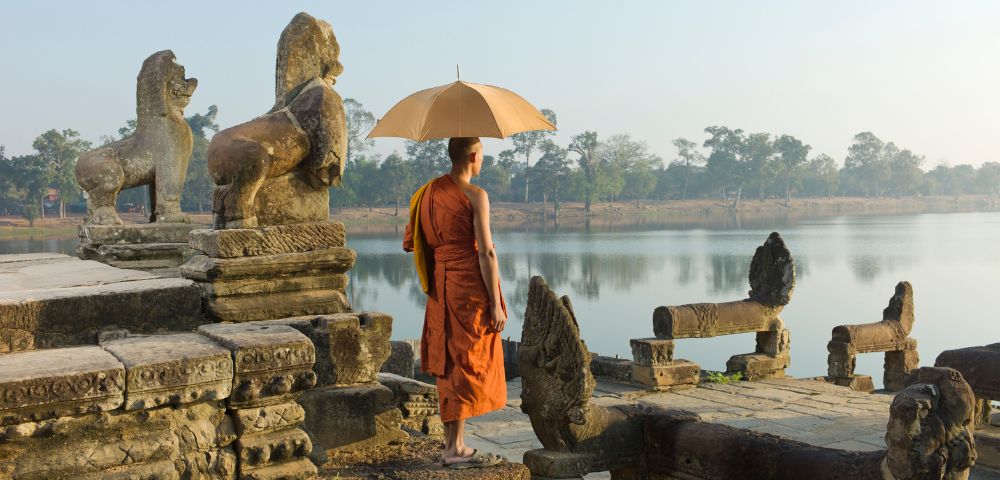 A monk in orange robes stands on ancient stone steps by a tranquil lake, holding a beige umbrella. Nearby are old lion statues. The mood is serene and reflective.