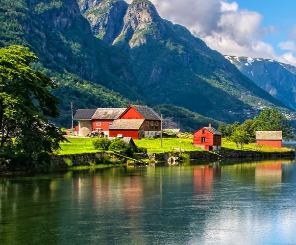 Photo 1 Continent: Europe Destination: Lofoten Islands, Norway Alt Text: Scenic view of Lofoten Islands with dramatic mountains, calm water, and colorful houses under a vibrant sky. Photo 2 Continent: Europe Destination: Norwegian Fjords, Norway Alt Text: Stunning fjord landscape with steep green cliffs, deep blue water, and a Norwegian flag in the foreground. Photo 3 Continent: Europe Destination: Norwegian Countryside, Norway Alt Text: Picturesque rural scene with red houses, lush greenery, and mountains reflected in a serene fjord.