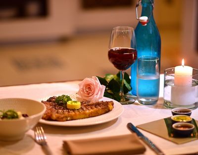 A romantic dinner setting with a grilled steak, a glass of red wine, a pink rose, and a lit candle. Warm ambiance with elegantly arranged tableware.