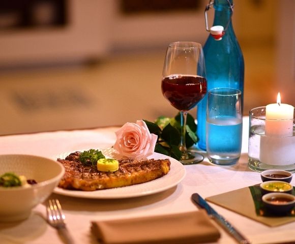 A romantic dinner setting with a grilled steak, a glass of red wine, a pink rose, and a lit candle. Warm ambiance with elegantly arranged tableware.