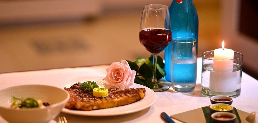 A romantic dinner setting with a grilled steak, a glass of red wine, a pink rose, and a lit candle. Warm ambiance with elegantly arranged tableware.