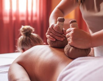 Massage therapy scene with a therapist applying herbal compresses to a person's back. The setting is serene, with warm, soft lighting and pink curtains.