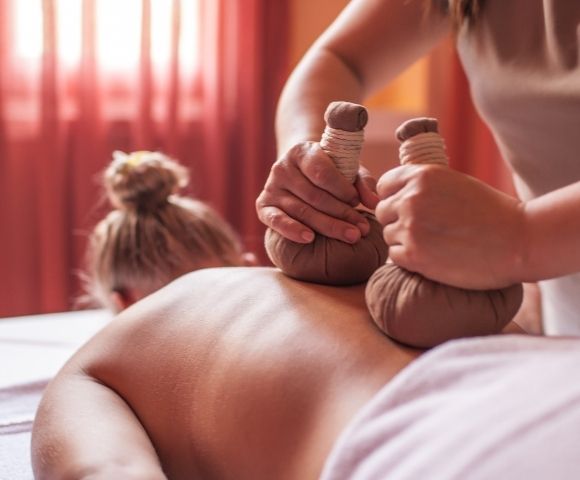 Massage therapy scene with a therapist applying herbal compresses to a person's back. The setting is serene, with warm, soft lighting and pink curtains.