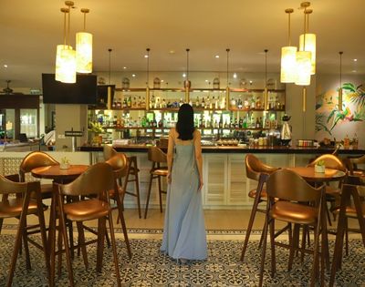 A woman in a long blue dress stands in a stylish bar with modern decor, warm lighting, and wooden furnishings, creating a cozy, elegant atmosphere.