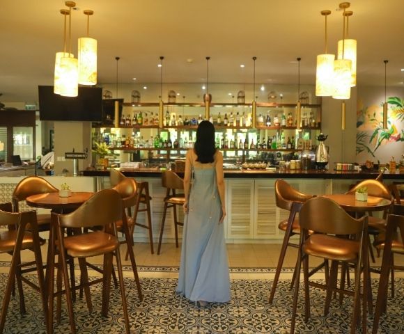 A woman in a long blue dress stands in a stylish bar with modern decor, warm lighting, and wooden furnishings, creating a cozy, elegant atmosphere.