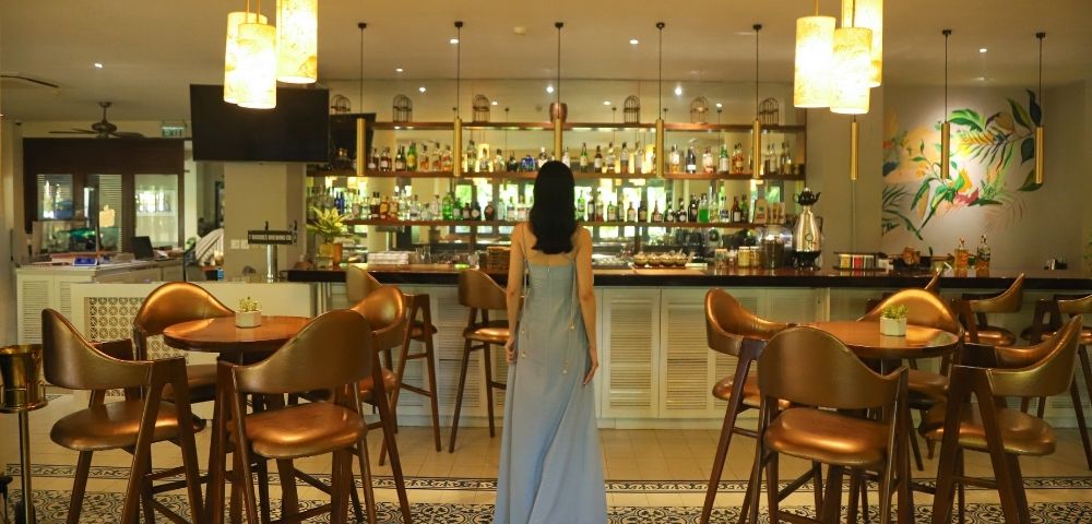 A woman in a long blue dress stands in a stylish bar with modern decor, warm lighting, and wooden furnishings, creating a cozy, elegant atmosphere.