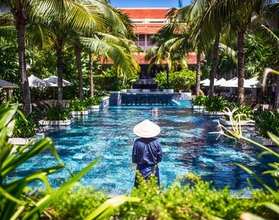 A person in traditional Asian attire and conical hat stands by a luxurious pool lined with palm trees on a sunny day, creating a serene tropical vibe.
