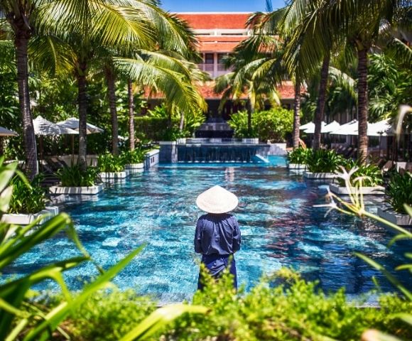 A person in traditional Asian attire and conical hat stands by a luxurious pool lined with palm trees on a sunny day, creating a serene tropical vibe.