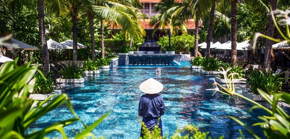 A person in traditional Asian attire and conical hat stands by a luxurious pool lined with palm trees on a sunny day, creating a serene tropical vibe.