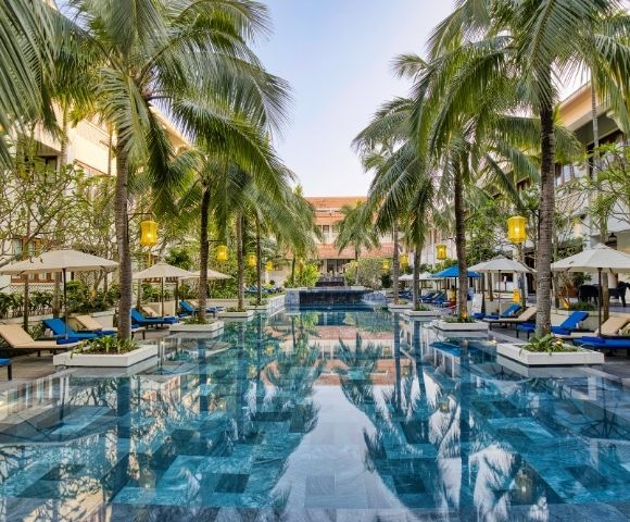 Luxurious hotel pool surrounded by tall palm trees and blue lounge chairs under umbrellas. The serene setting reflects a clear blue sky.