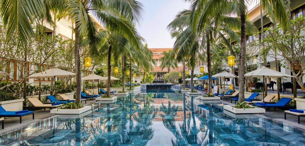 Luxurious hotel pool surrounded by tall palm trees and blue lounge chairs under umbrellas. The serene setting reflects a clear blue sky.