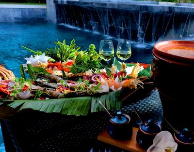 A vibrant platter of seafood and vegetables on banana leaves by a poolside, with two wine glasses. It's a serene setting with an elegant and refreshing ambiance.
