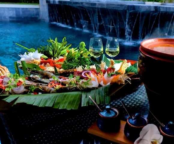 A vibrant platter of seafood and vegetables on banana leaves by a poolside, with two wine glasses. It's a serene setting with an elegant and refreshing ambiance.