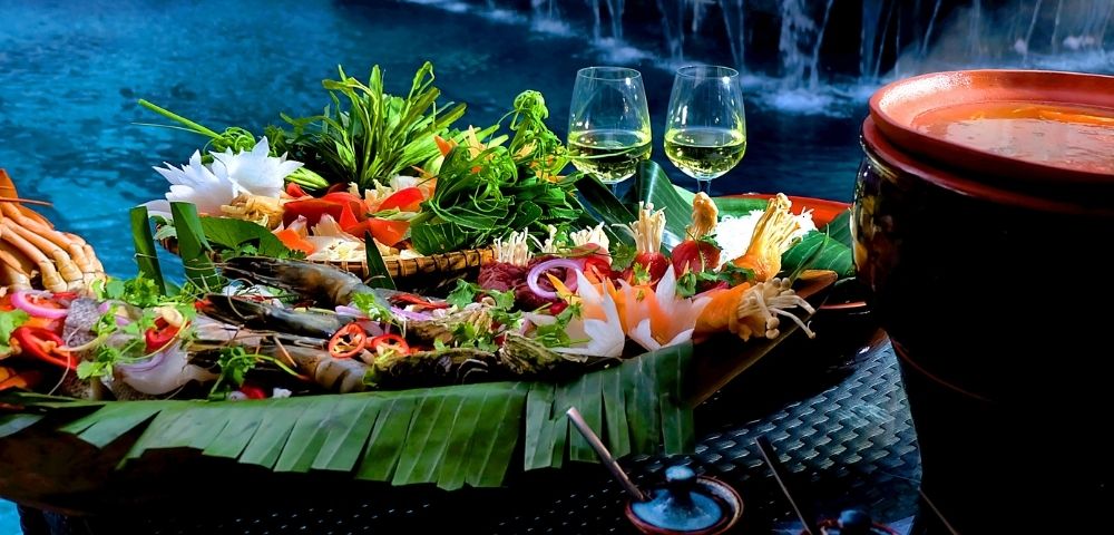 A vibrant platter of seafood and vegetables on banana leaves by a poolside, with two wine glasses. It's a serene setting with an elegant and refreshing ambiance.
