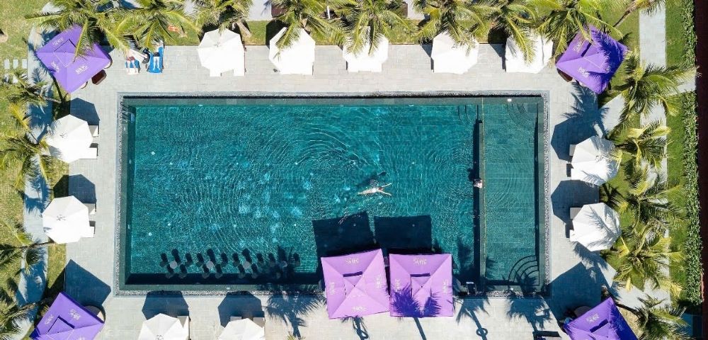 Aerial view of a serene swimming pool surrounded by palm trees and white umbrellas, with a person swimming. Purple canopies provide a vibrant contrast.