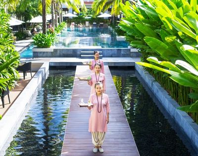 Three women in pink dresses walk along a wooden path over a tranquil pond lined with lush greenery, leading to a serene pool. The scene is peaceful and elegant.