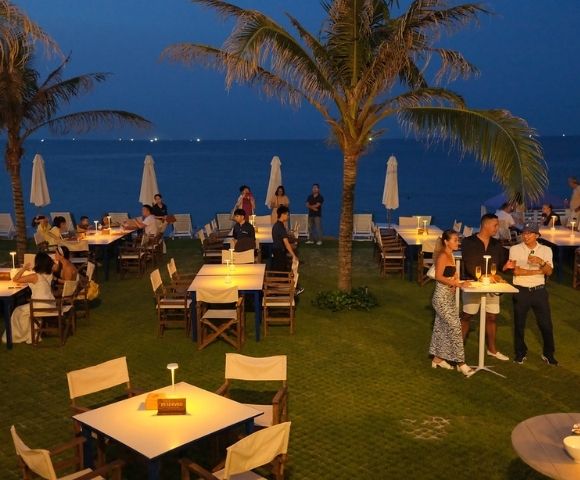 Outdoor restaurant by the sea at evening, with dimly lit tables, palm trees, patrons dining and socializing. A calm, tropical ambiance is conveyed.