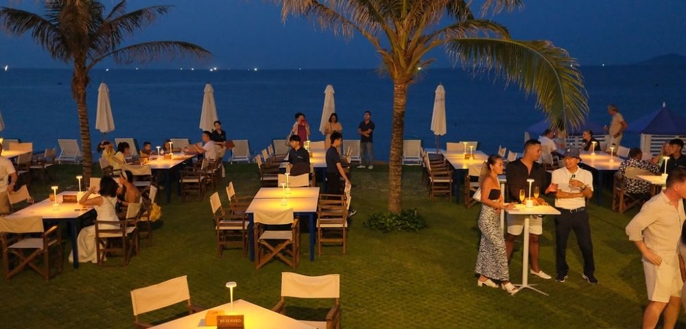 Outdoor restaurant by the sea at evening, with dimly lit tables, palm trees, patrons dining and socializing. A calm, tropical ambiance is conveyed.
