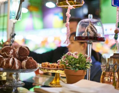 Pastry display in a vibrant market setting. Foreground includes decorative desserts, potted plant, and utensils. Blurred background with soft lighting.
