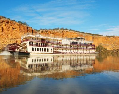 A large paddle steamboat with red and white accents sails on calm water, reflecting the boat and surrounding golden cliffs under a clear blue sky.