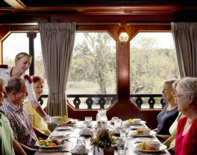 A group of five people dine on a boat, with a server attending them. The table is set with dishes, and large windows offer a view of trees outside. Cozy atmosphere.