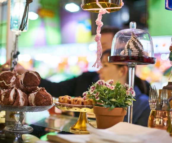 Pastry display in a vibrant market setting. Foreground includes decorative desserts, potted plant, and utensils. Blurred background with soft lighting.
