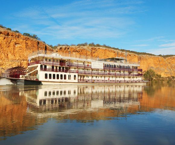A large paddle steamboat with red and white accents sails on calm water, reflecting the boat and surrounding golden cliffs under a clear blue sky.