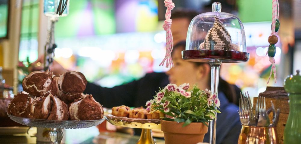Pastry display in a vibrant market setting. Foreground includes decorative desserts, potted plant, and utensils. Blurred background with soft lighting.