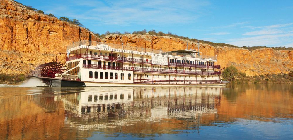 A large paddle steamboat with red and white accents sails on calm water, reflecting the boat and surrounding golden cliffs under a clear blue sky.