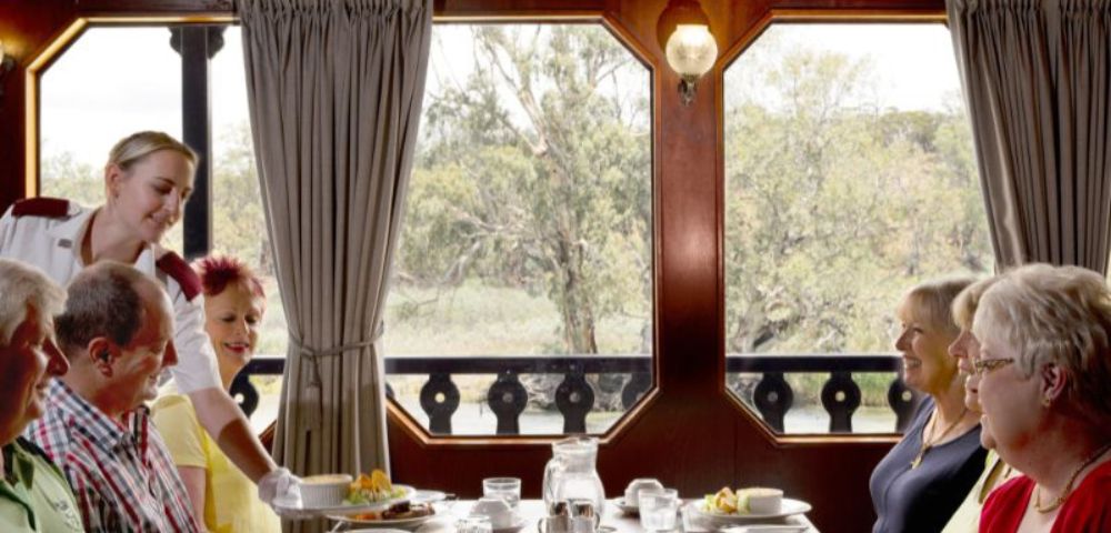 A group of five people dine on a boat, with a server attending them. The table is set with dishes, and large windows offer a view of trees outside. Cozy atmosphere.