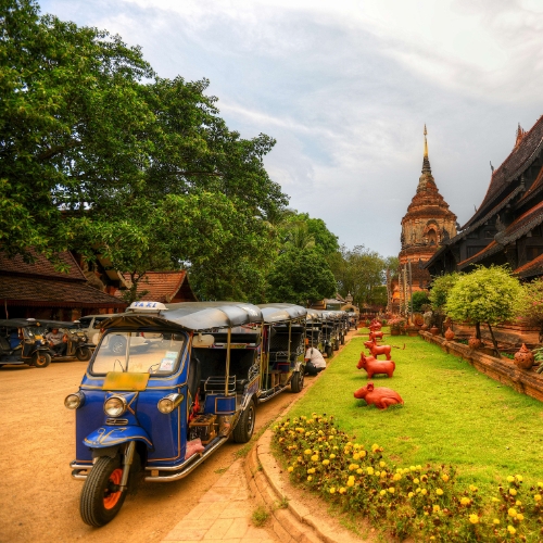 Tuk-tuks lined up on a dirt path beside a lush park with red statues and yellow flowers. A temple spire rises in the background, under a cloudy sky.
