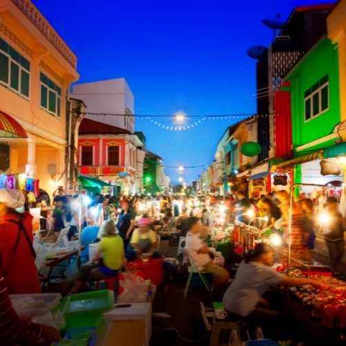 A vibrant night market scene with colorful buildings and bright lights. People browse stalls selling various goods, creating a lively, bustling atmosphere.