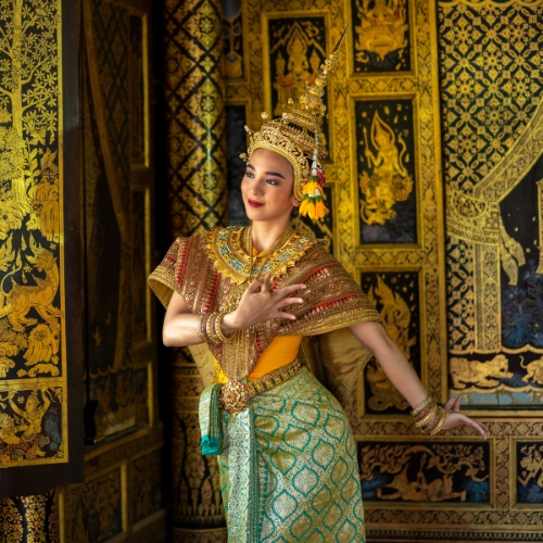 A dancer in traditional Thai attire poses against ornate golden murals. The dancer wears a gold headdress and colorful costume, exuding grace and elegance.
