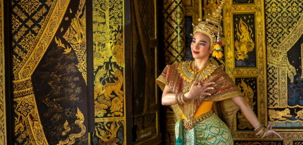 A dancer in traditional Thai attire poses against ornate golden murals. The dancer wears a gold headdress and colorful costume, exuding grace and elegance.