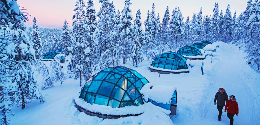 Two people stroll through a snowy forest, surrounded by unique glass domes glistening in the winter sunlight.