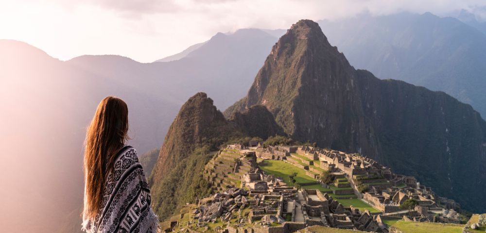 A woman in a traditional dress stands at the edge of Machu Picchu, overlooking the ancient ruins and lush mountains.