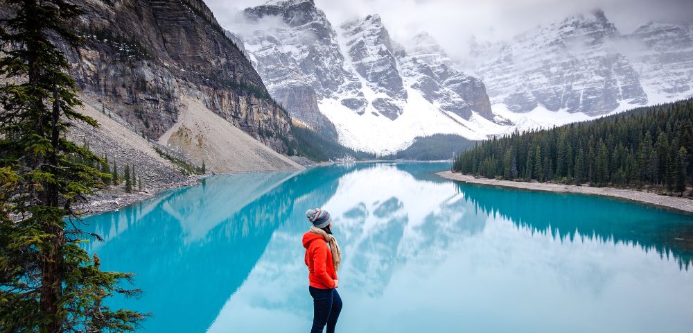 A woman stands on a rock, gazing out over a serene lake surrounded by trees and mountains.