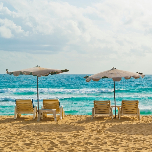 Four loungers and two umbrellas on a sandy beach overlook a turquoise ocean under a partly cloudy sky, evoking a relaxed, serene mood.
