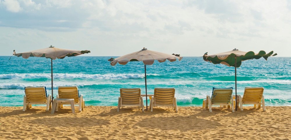 Six loungers and three umbrellas on a sandy beach overlook a turquoise ocean under a partly cloudy sky, evoking a relaxed, serene mood.