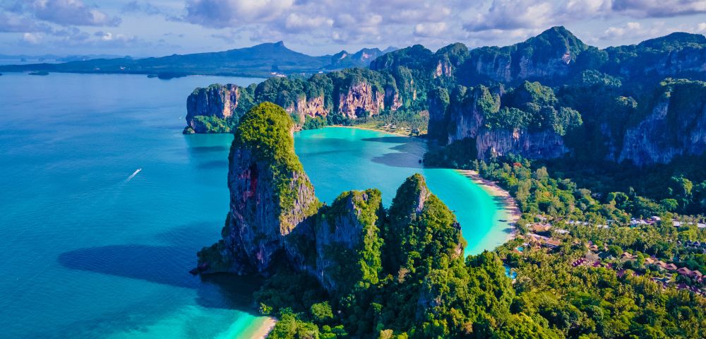 Aerial view of a tropical coastline with turquoise waters, lush greenery, and steep cliffs under a sunny blue sky. The tranquil scene evokes a sense of serenity.