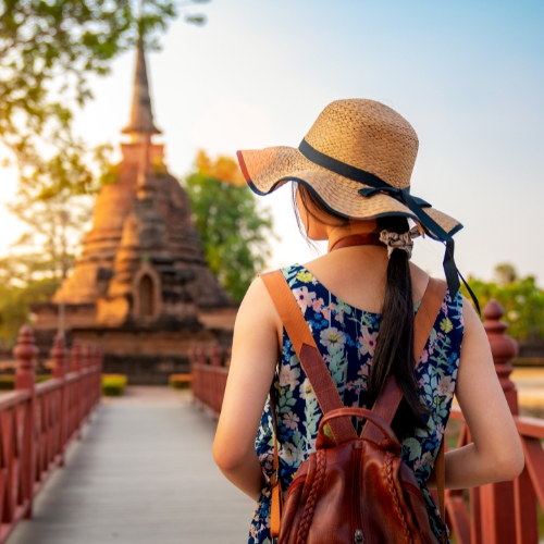 A woman in a floral dress and sunhat stands on a bridge, facing a historic temple in the distance. The scene conveys warmth and exploration.