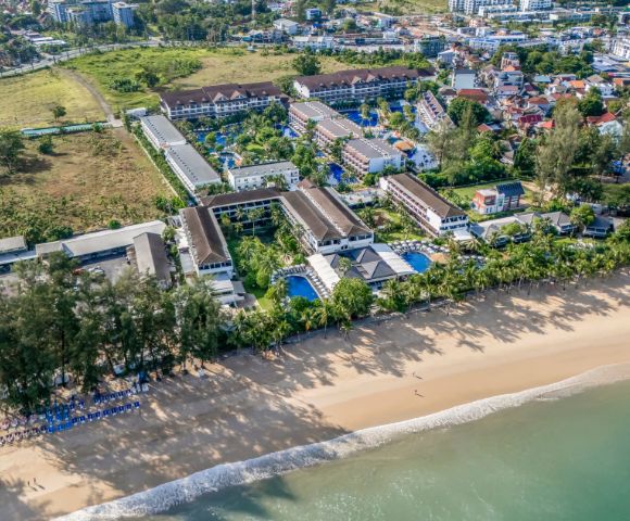 Aerial view of a coastal resort with lush trees, pool-lined buildings, and a sandy beach. The vibrant blue sea exudes tranquility and warmth.