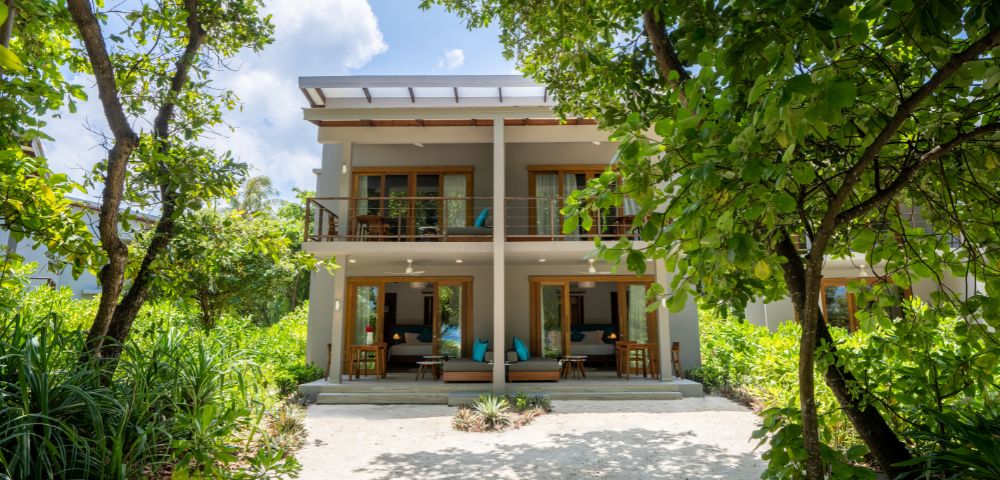 A modern two-story villa surrounded by lush greenery, featuring large windows and balconies with outdoor seating. Blue skies above.