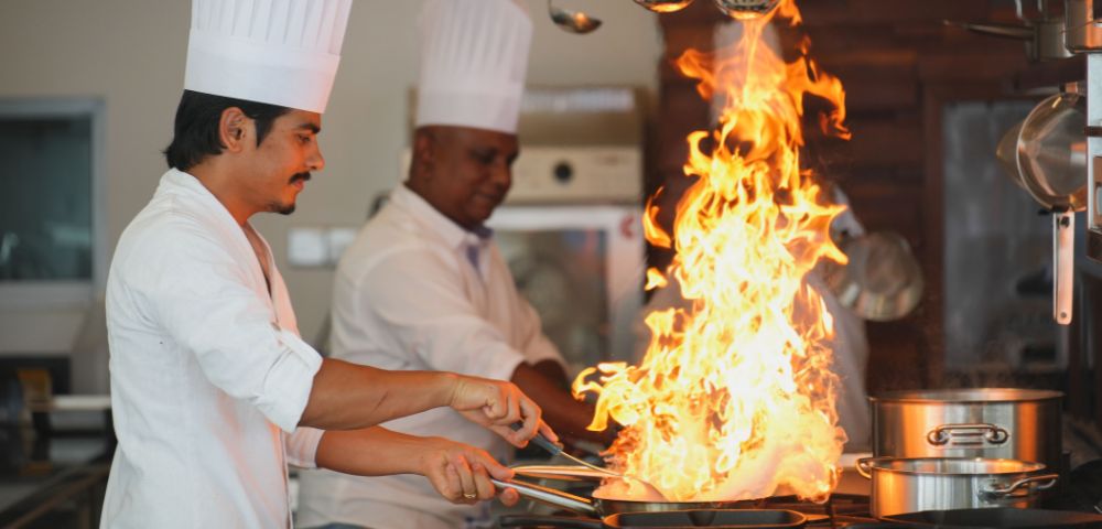 Two chefs in white uniforms are cooking in a kitchen, with flames erupting from a frying pan, creating a dramatic cooking scene.
