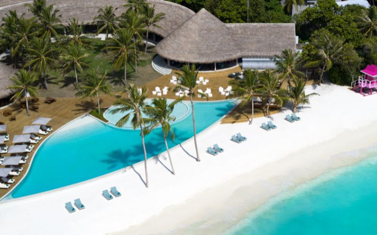 Aerial view of a resort featuring a sparkling pool and a sandy beach, surrounded by lush greenery.