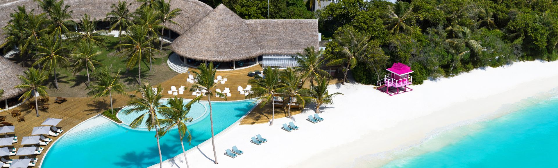 Aerial view of a resort featuring a sparkling pool and a sandy beach, surrounded by lush greenery.