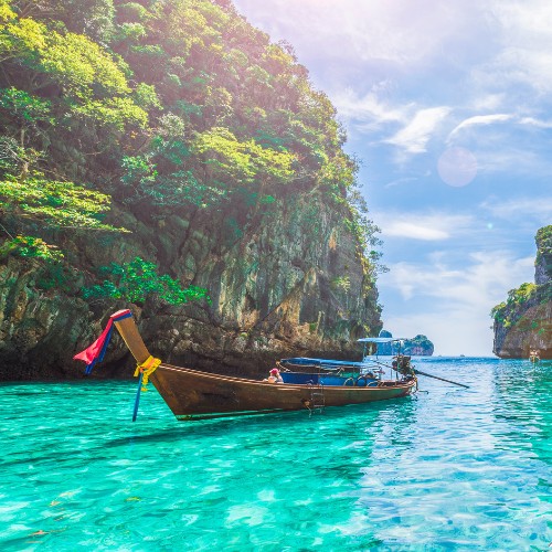 Long-tail boat floats on clear turquoise water, surrounded by lush green cliffs under a sunny, partly cloudy sky, conveying a serene, tropical feel.