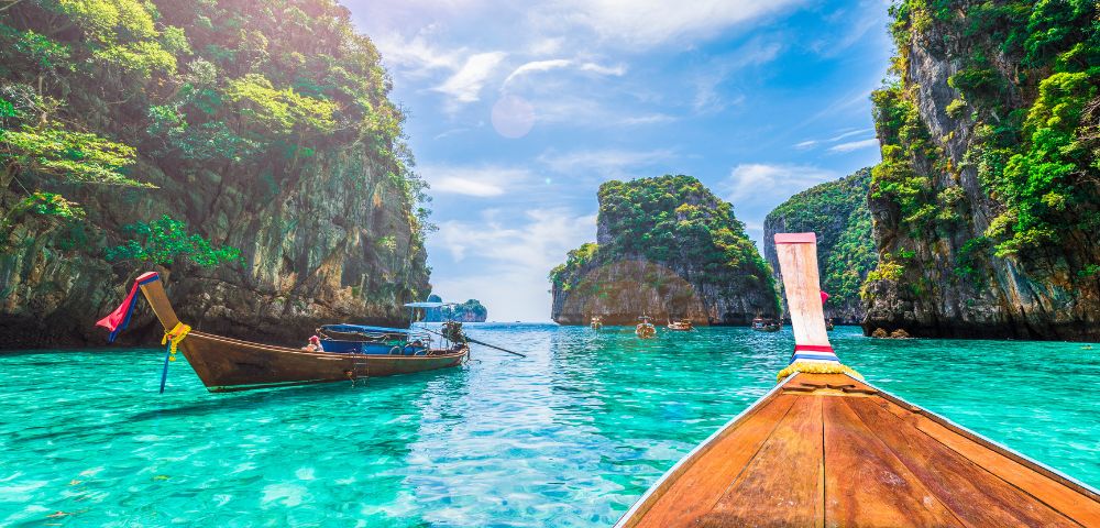 Turquoise waters surround wooden boats between lush, towering cliffs under a bright blue sky. The scene exudes tranquility and tropical beauty.