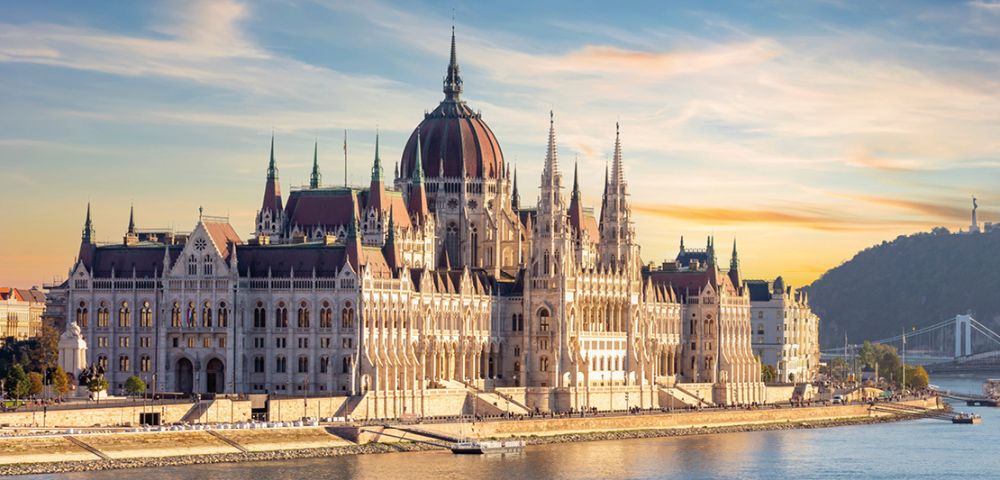 A grand Gothic-style building with a central dome and spires, bathed in warm sunset light, overlooks a calm river. The sky is clear with soft clouds.