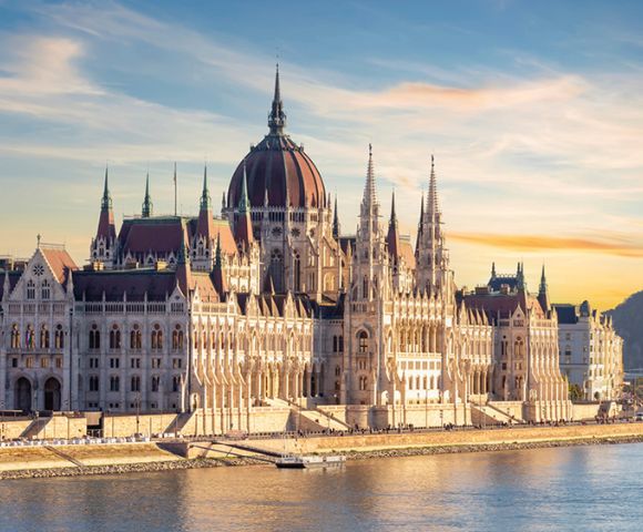 A grand Gothic-style building with a central dome and spires, bathed in warm sunset light, overlooks a calm river. The sky is clear with soft clouds.